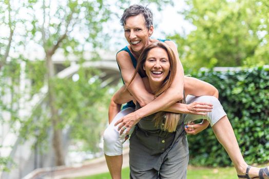 Mature woman giving piggyback to mature friend female in the park. Middle aged friends sharing time together and having fun.