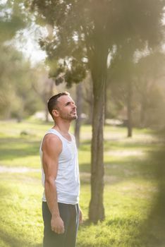 Man wearing white sleeveless t-shirt looking away in the park. Young adult standing in the park with arms close to his body looking up.