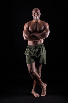 African American man standing looking at camera with arms crossed and one leg crossed. Muscular adult male wearing only shorts in a studio with black background.