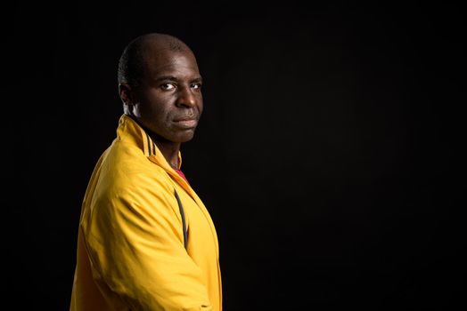 Side view of an African American man standing looking at camera. Adult male in yellow jacket and red t-shirt in a studio with black background.