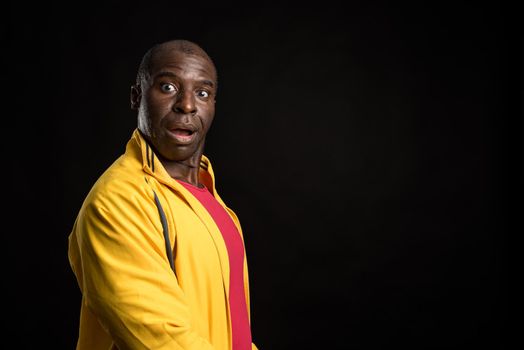 Side view of an African American man standing looking at camera. Adult male in yellow jacket and red t-shirt in a studio with black background.