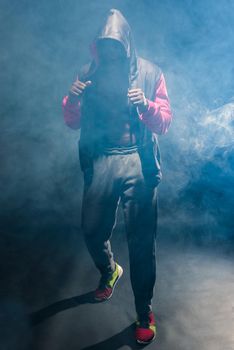 Muscular adult male in hooded jacket with bare torso with smoke around standing in defensive boxing pose. African American man in sportswear standing in a studio with black background.