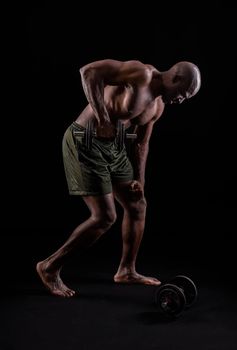 Side view of a muscular man standing leaning forward doing triceps exercise with dumbbells on a black background. African american man with bare torso doing exercise for arms.