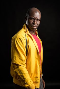 Side view of an African American man standing looking distrustful . Adult male in yellow jacket and red t-shirt in a studio with black background.