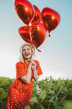 Portrait of attractive woman in red dress posing with heart-shaped balloons on green nature background. Girl in straw vintage hat or beret. Birthday, holiday, celebrate freedom concept. High quality