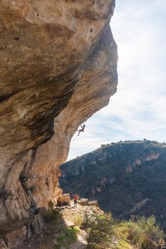 Distant view of a rock climber hanging and climber a rock formation. Vertical view of a bare torso climber climbing a rock with a background of hill and sky.