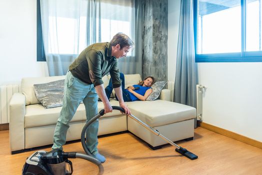 Man vacuuming the living room while his wife is lying on the couch. High quality photo