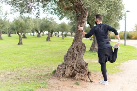 Young runner man with mask stretching in a park leaning against a tree. High quality photo