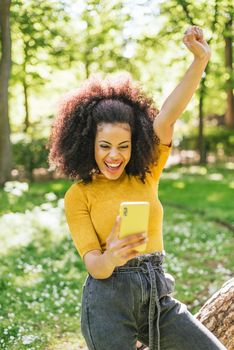 Pretty afro woman chatting on her mobile, celebrating good news with her fist raised. In a park. Selective focus.