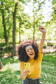 Pretty afro woman chatting on her mobile, celebrating good news with her fist raised. In a park. Selective focus.