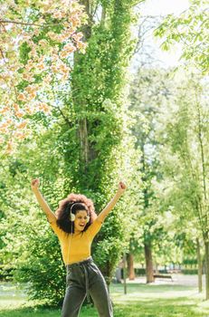 Pretty afro woman listens to music with headphones dancing in a garden. Selective focus.