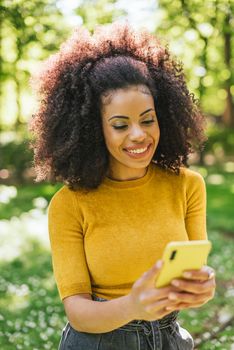 Pretty afro woman typing on mobile, in the forest. Selective focus.