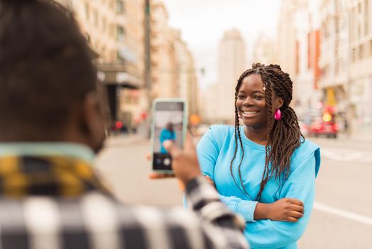 Black guy taking a photo with his mobile of his girlfriend in the middle of the city. Selective focus.