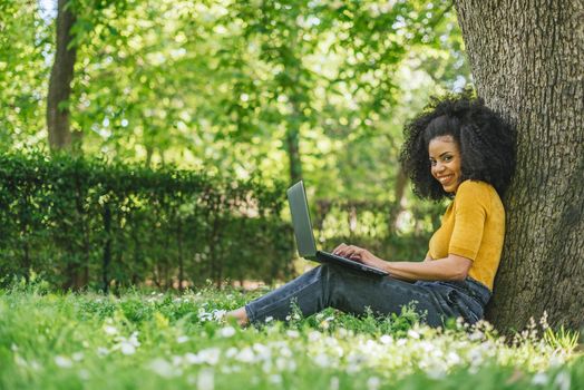 Beautiful and happy afro woman typing on a laptop in a garden. Side view at grass level.