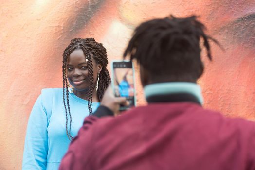 Black guy taking a photo with his mobile of his girlfriend in the middle of the city. Selective focus.