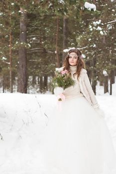 Beautiful bride in a white dress with a bouquet in a snow-covered winter forest. Portrait of the bride in nature.