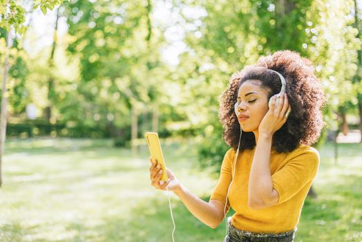 Pretty afro woman listens to music with headphones dancing in a garden. Selective focus.