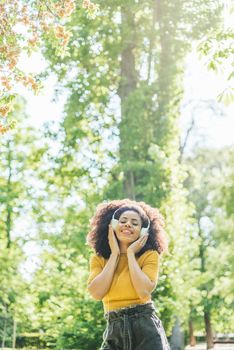 Pretty afro woman listens to music with headphones dancing in a garden. Selective focus.