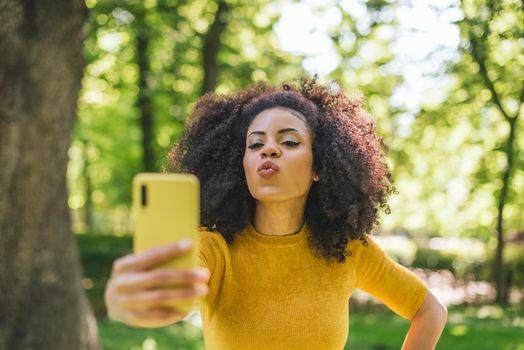 Pretty afro woman taking a selfie blowing a kiss, in the forest. Selective focus.