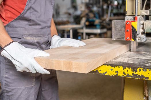 Crop anonymous male artisan in protective wear cutting timber board with sharp blade of band saw in professional joinery factory