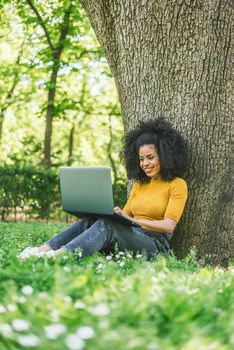 Beautiful and happy afro woman typing on a laptop in a garden. Side view at grass level.