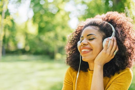 Pretty afro woman listens to music with headphones dancing in a garden. Selective focus.