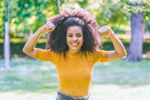 Portrait of pretty afro girl in a garden, pulling out biceps muscle. Selective focus.