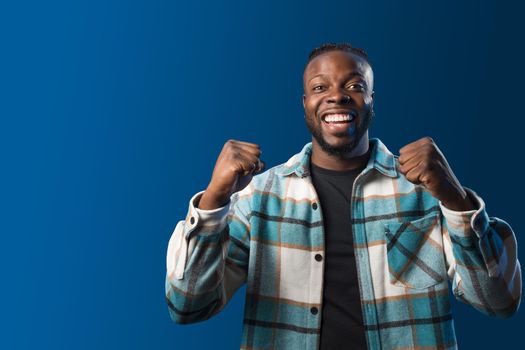 Handsome black man smiling, celebrating something with his fists raised. Blue background