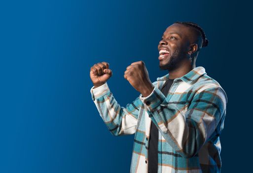 Handsome black man smiling, celebrating something with his fists raised. Mid shot. Blue background