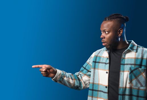 Handsome black man pointing aside, surprised. Mid shot. Blue background.