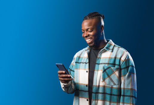 Handsome black man looking at mobile smiling. Mid shot. Blue background.