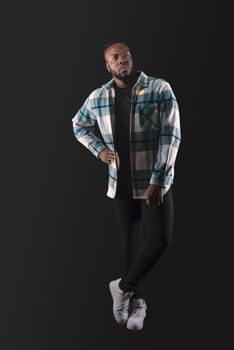 Handsome black man, standing looking serious to the side, wearing blue plaid shirt and jeans, legs crossed. Full body. Blue background.