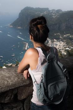 Pretty woman from behind with sunglasses and backpack facing the sea from the top on a sunny summer day. Capri island.