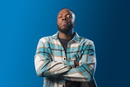 Handsome black man, serious and angry, crossed arms. Mid shot. Blue background.