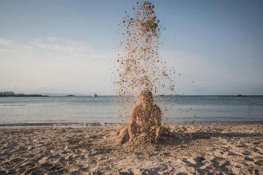 Man kneeling on a beach throwing sand at himself. Formentera island, Spain.