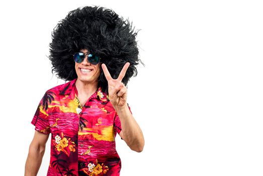 Happy man in vibrant shirt with sunglasses and funny Afro wig looking at camera with smile and showing V sign gesture against white background