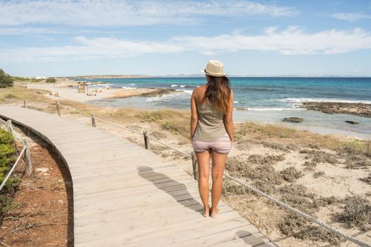 Woman in a T-shirt and hat, standing looking out to sea from a little path made of wooden boards. Long shot. Formentera island, Spain.