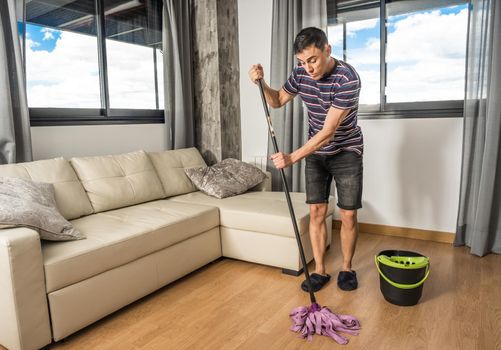 Man in casual clothes and slippers mopping the living room floor. Full body.