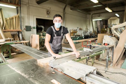 Worker with mask and protective equipment cutting on a sliding table saw in a factory. Mid shot.