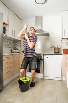 Man in the kitchen wearing casual clothes and slippers, wringing the mop into the bucket with excessive force. Full body.