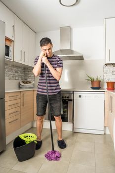 Sad and pensive man wearing casual clothes and slippers mopping the kitchen floor. Full body.