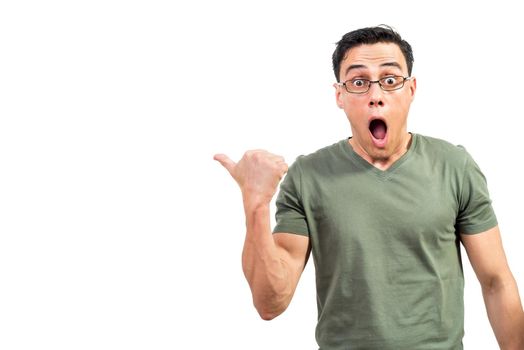 Astonished male nerd in t shirt and glasses looking at camera with opened mouth and pointing away with thumb against white background