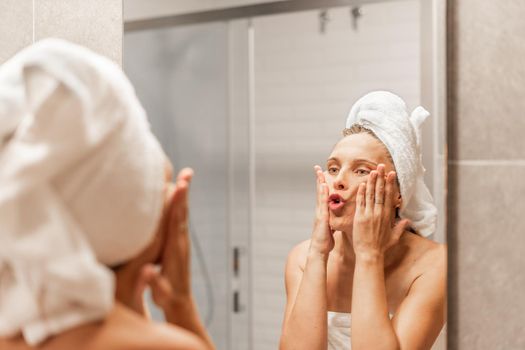 Funny caucasian adult woman putting moisturizer on her face with both hands and massaging her face while looking at herself in a hotel mirror. Concept of skin care.