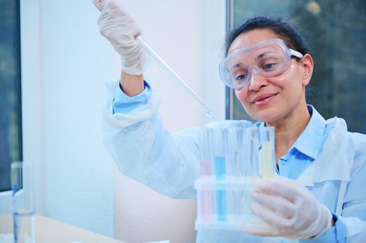Inspired Latin American woman, lab assistant, scientist chemist using a glass dropper, dripping few chemical reagent into test tubes, while conducting experiment in a clinical research laboratory