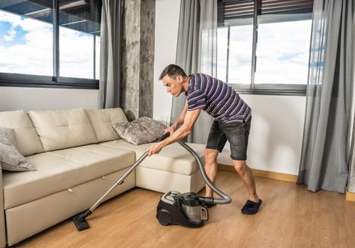 Man with casual wear, vacuuming under the living room sofa. Full body.