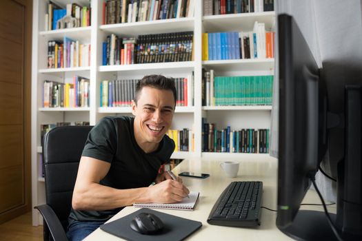Smiling and happy man in his room sitting at the computer looking at camera, wearing a shirt. Mid shot.