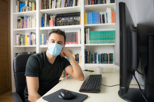 Man teleworking at home, sitting in front of the computer wearing a shirt and mask. Mid shot.