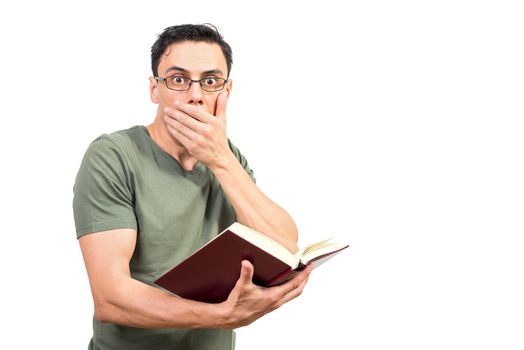 Male nerd in glasses covering mouth and looking at camera in astonishment while reading book isolated on white background