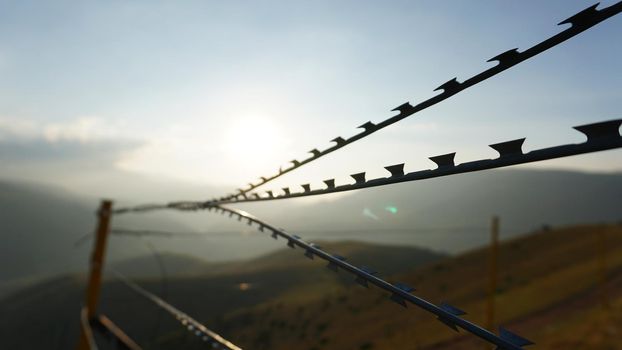 Sharp barbed wire on the fence with a blurred background of mountains. A bright yellow sun in a blue sky. Gray clouds. The high hills are covered with yellow-green grass. Restricted area. Background