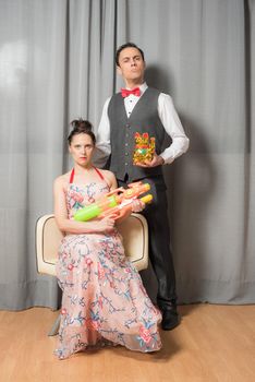 Eccentric marriage portrait with solemn attitude. The woman, seated in an elegant flowered dress, carrying a toy weapon. The man, standing, wearing an elegant suit and carrying a toy kitten.